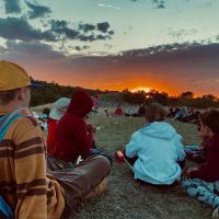 A group of young people sit watching a brilliant, colourful sunset.