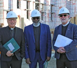 Three ministers wearing clerical collars and hard hats stand in front of a construction site