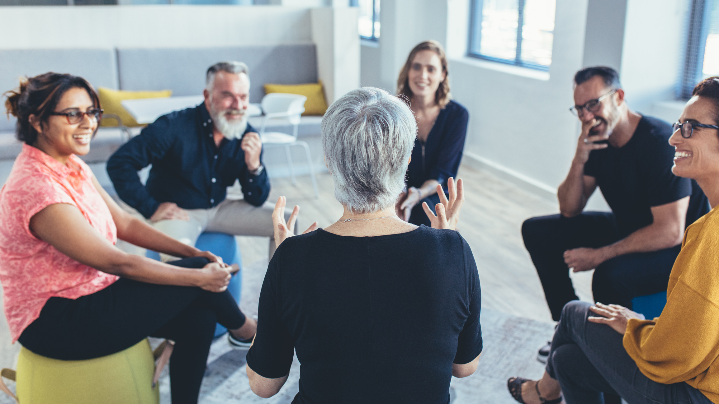 Six people sit in a circle in a room, five of them listening to a woman who is talking.