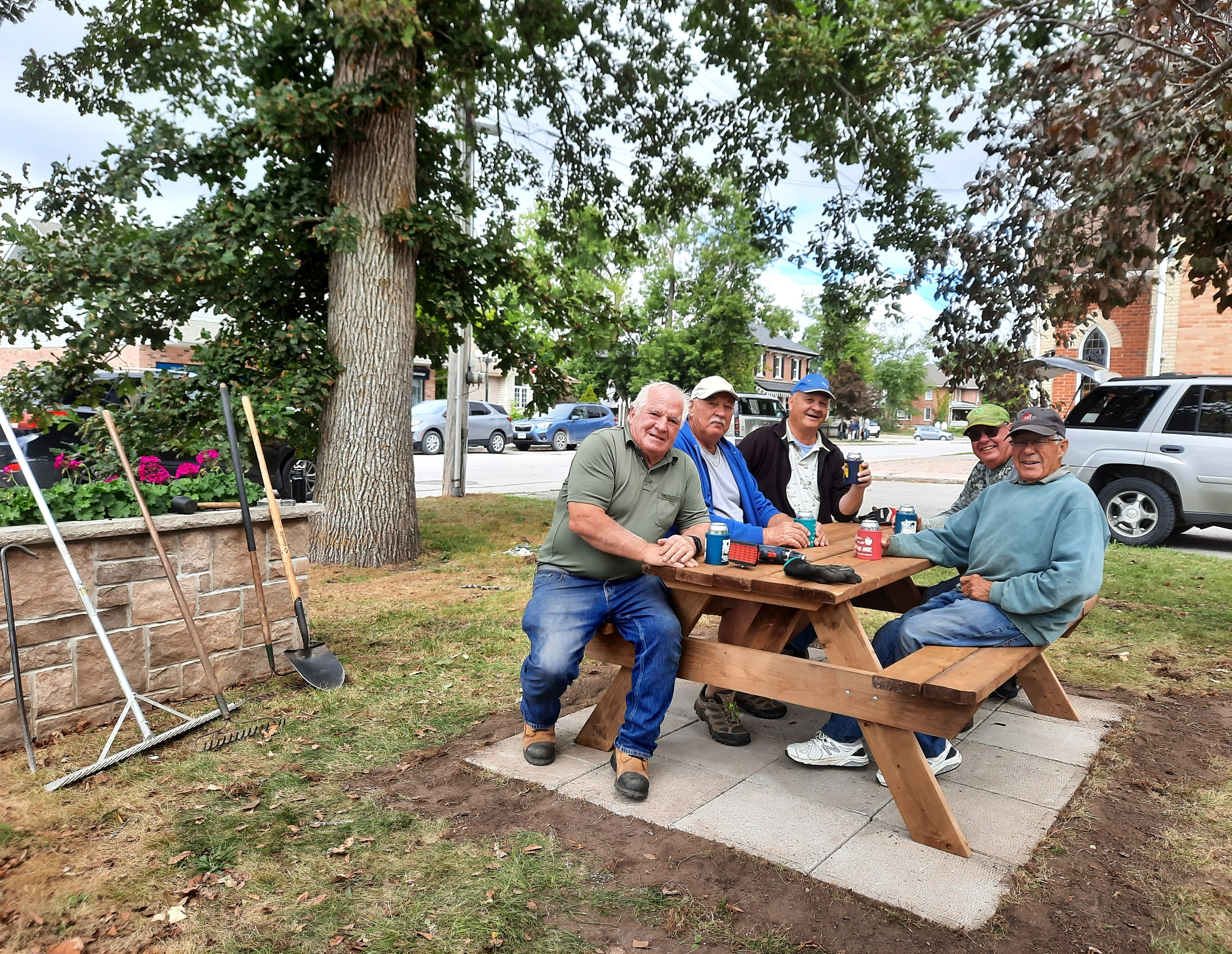 Five men sitting at a picnic table drinking coffee under a tree