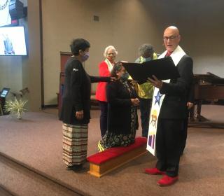 Deb Anderson-Pratt kneels at ordination ceremony while minister reads and woman presents her with eagle feather