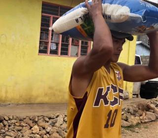 A man carries a large bag of rice and a yellow house is in the background.