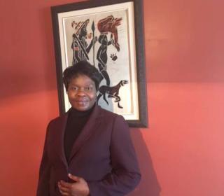 The Rev. Marie-Claude Manga, standing in front of an artwork, dressed in a brown jacket.