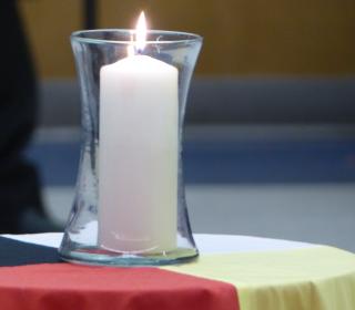 Burning Tall white candle in clear glass jar sits on a cloth with the four Indigenous colours of black, white, yellow, and red.