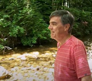 Alf Dumont, an Indigenous man, stands beside a creek, smiling and looking upstream.