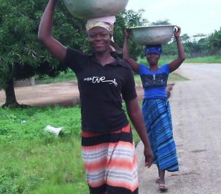 Two Zambian women walk along a road carrying large metal tubs on their heads.