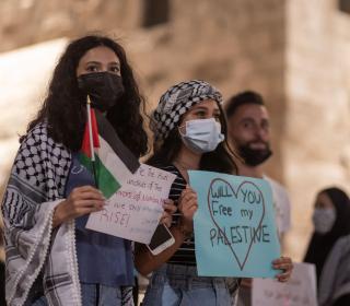 Two Palestinian women hold up candles and signs at a protest. One of the signs reads Will You Free My Palestine.