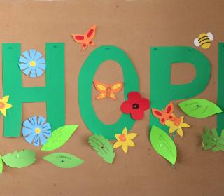 The word "hope" spelled out on wall in green paper letters surrounded by paper flowers, leaves with notes written on them, and insects.