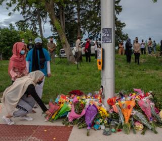 Two women and a man lay flowers at a makeshift sidewalk memorial while several people look on.