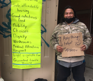 A man with dark skin and rough clothing holding a sign that says "Anything helps," next to another homemade sign listing the benefits of a Guaranteed Livable Income.