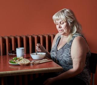 A woman sits at a table eating soup, bread, and a salad against a brown wall.