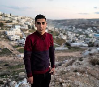 Image: A teenage Palestinian boy wearing a maroon sweater standing in front of a hilly and rocky background.