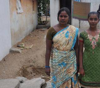 An Indian woman and her teenage daughter stand in front of a simple concrete house.