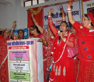 Seven Indian women dressed in red stand at a podium with their right arms raised. 