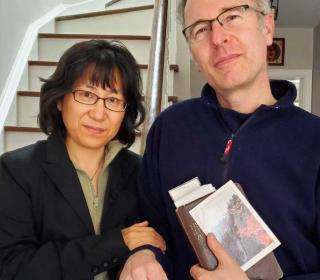 HyeRan Kim-Cragg (left) and husband David Kim-Cragg (right), stand at the foot of a staircase in a house. David is holding a Bible and a postcard of the Diamond Mountains in Korea. Both are wearing black.