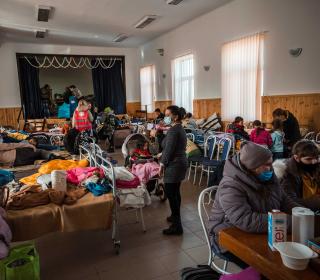 In a crowded refugee centre people sit or stand around beds lined up along the walls.