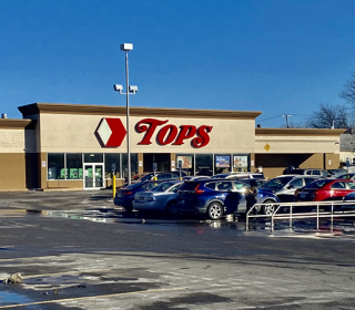 Exterior of a supermarket with the name Tops in large red letters over the doors and cars parked in front. 