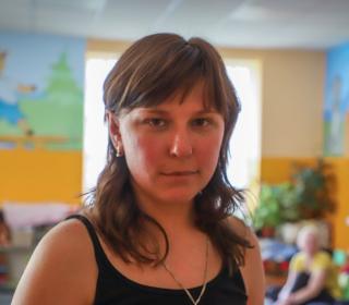 A woman in her 30s looks into the camera. In the background we see a room decorated with children's art and toys.