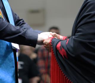 The hands of someone wearing a minister's stole and another person wearing an Indigenous shawl are clasped. 