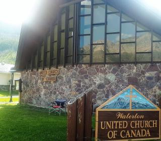Exterior shot of a church seen against a mountain with a sign in front saying Waterton United Church