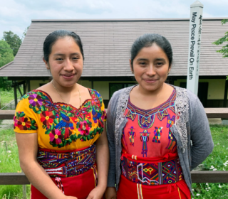 Two young women dressed in Indigenous Guatemalan clothing stand in front of a building in Japan. 