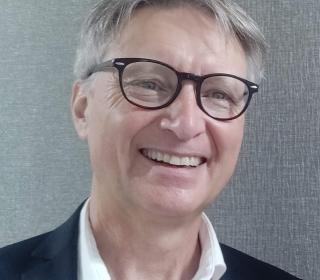 A portrait of Rob Dalgleish, executive director of EDGE Network for Ministry Development. He is a white man with greying hair, large glasses, and an enthusiastic smile. Wearing an open neck white dress shirt and jacket.