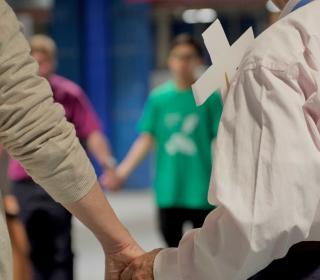 Two hands hold each other, part of a circle of people seen in the background.