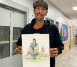A man wearing a black hoodie and baseball cap stands in a hallway holding his drawing of Indigenous people.