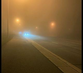 A moody photograph of a foggy street at night, with street lights dimly showing through and the headlights of a car approaching in the darkness.