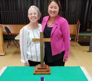 Rev. Leigh Sinclair (left) and Rev. MiYeon Lim (right) smile together as they stand before a temporary altar with a wooden cross and a small candle.