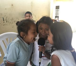 Three laughing little girls crowd around a microphone one is holding while a little boy looks on.