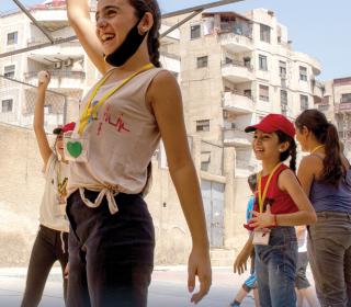 Cover of the 2022-2023 Gifts with Vision print catalogue showing girls in Damascus playing a game outdoors with highrise apartment buildings in the background.