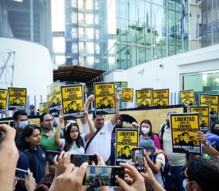 A crowd of people hold up yellow and black signs saying Libertad por los lideres comunitarios in front of an office building.