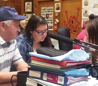An older man and two young women sit at a desk stacked with papers, books, and binders. In the background are more small groups sitting at tables. 