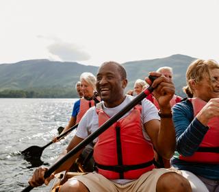 Six adults wearing life jackets paddle a canoe in a lake with mountains in the background.