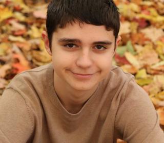 A White teenager with short dark hair sits in fall leaves and looks at the camera with a slight smile.
