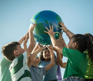 A diverse group of children, outside in green shirts, work together to hold up a ball decorated to look like Earth.