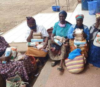 Five older African women are seated on the porch of a building holding parcels and shopping bags.