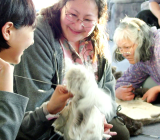 A young woman sews fur while an older woman looks on smiling