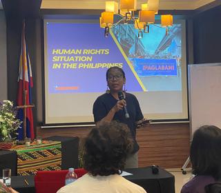 A woman stands in front of a group speaking into a microphone. Behind her a screen displays a PowerPoint slide saying Human rights situations in the Philippines.