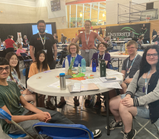 Several young people sit around a table in a gym where a big meeting is happening.