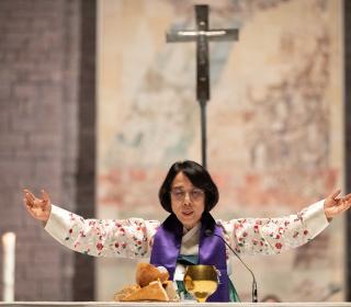 Rev. EunKyung Kim holds her arms out above a communion chalice and bread. A cross hangs on the wall behind her.