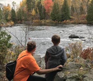 An adult woman crouches down next to a young child as they observe a flowing river, surrounded by autumn trees.