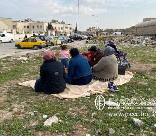 A group of women sit on a blanket in the middle of rubble