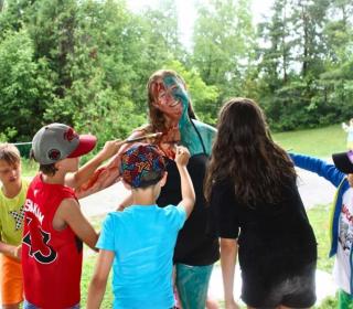 Children surround a camp counsellor with paintbrushes, painting her.