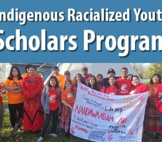 A large group of young people in orange shirts stand in front of a tepee hold a banner with Indigenous words on it