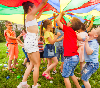 A group of children hold a large rainbow-coloured sail over them.