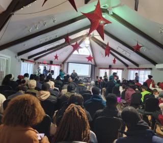 Young people fill a large room watching a presentation at the front.