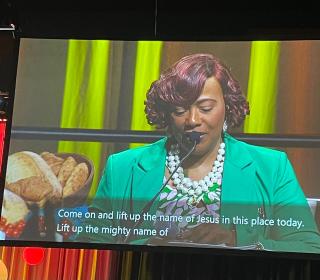 A photo of a woman speaking into a microphone projected onto a large screen