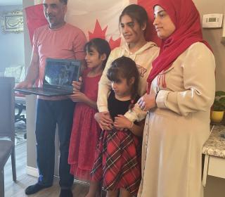 A family of two parents and three young girls holding up a laptop during an online citizenship ceremony.